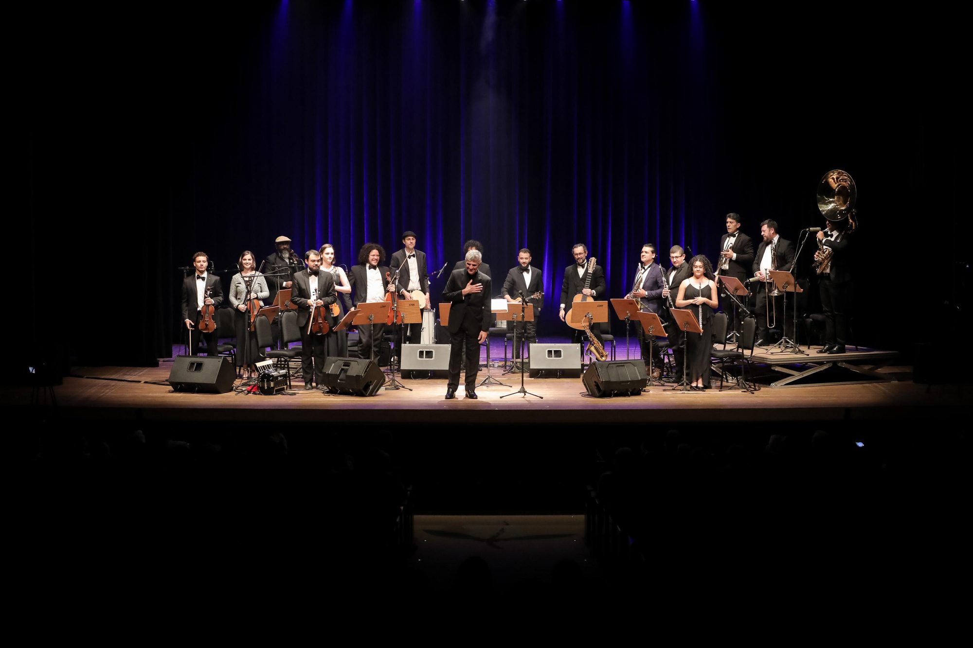 A orquestra popular do Clube do Choro agradece de pé no palco do Cine Teatro Ouro Verde, com o maestro à frente