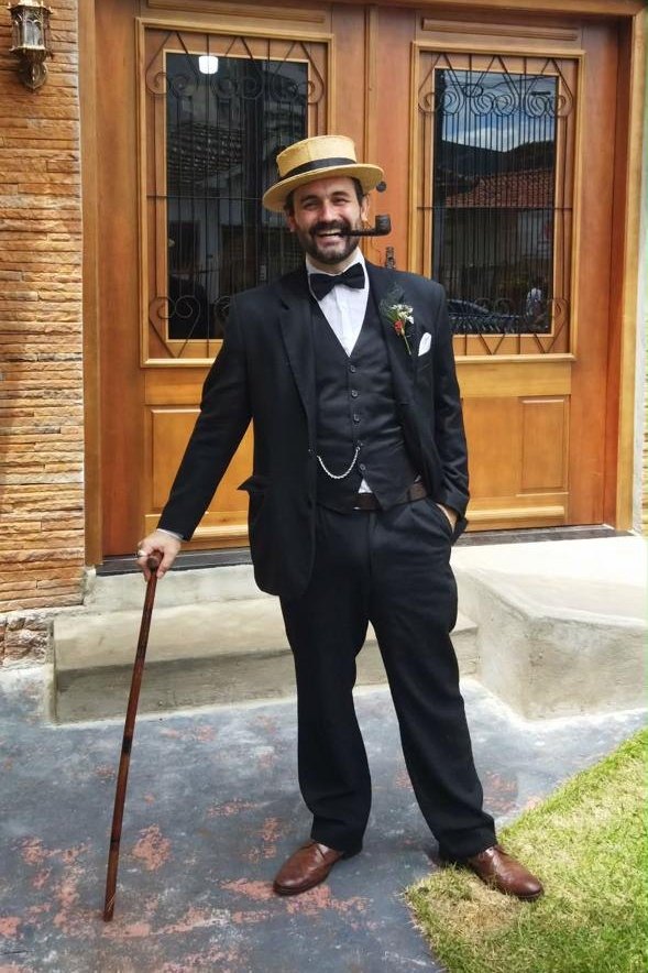 Osório Perez de terno preto, chapéu de palha, bengala e cachimbo, diante de uma porta de madeira