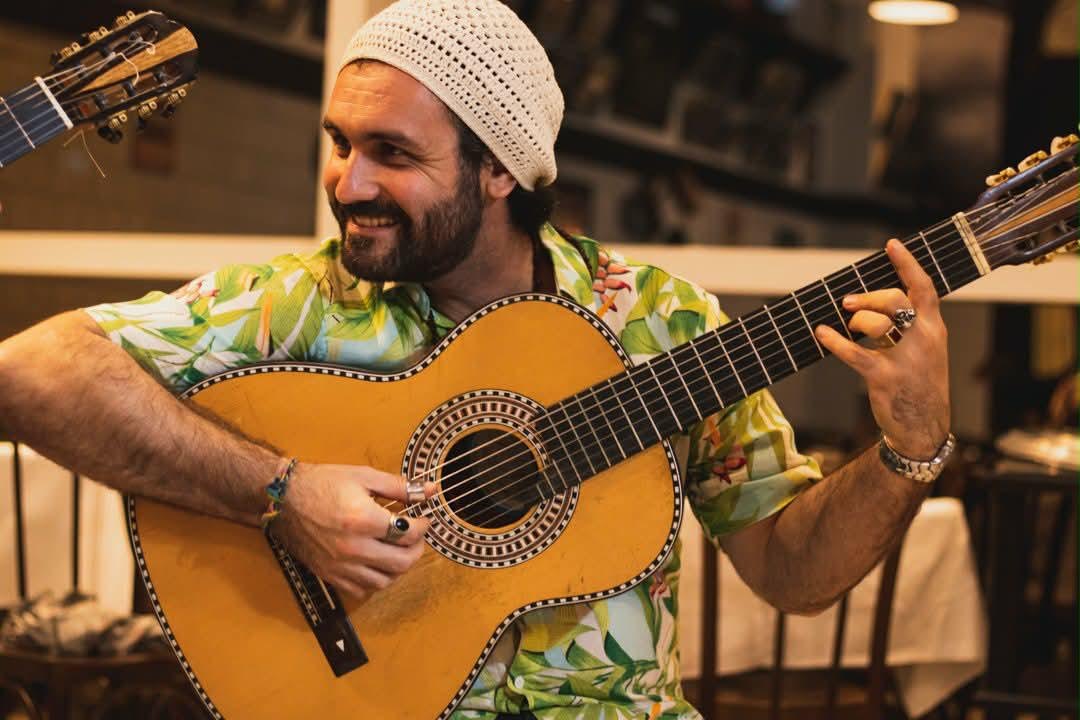Osório Perez sorrindo, de gorro branco e camisa estampada, tocando violão numa roda