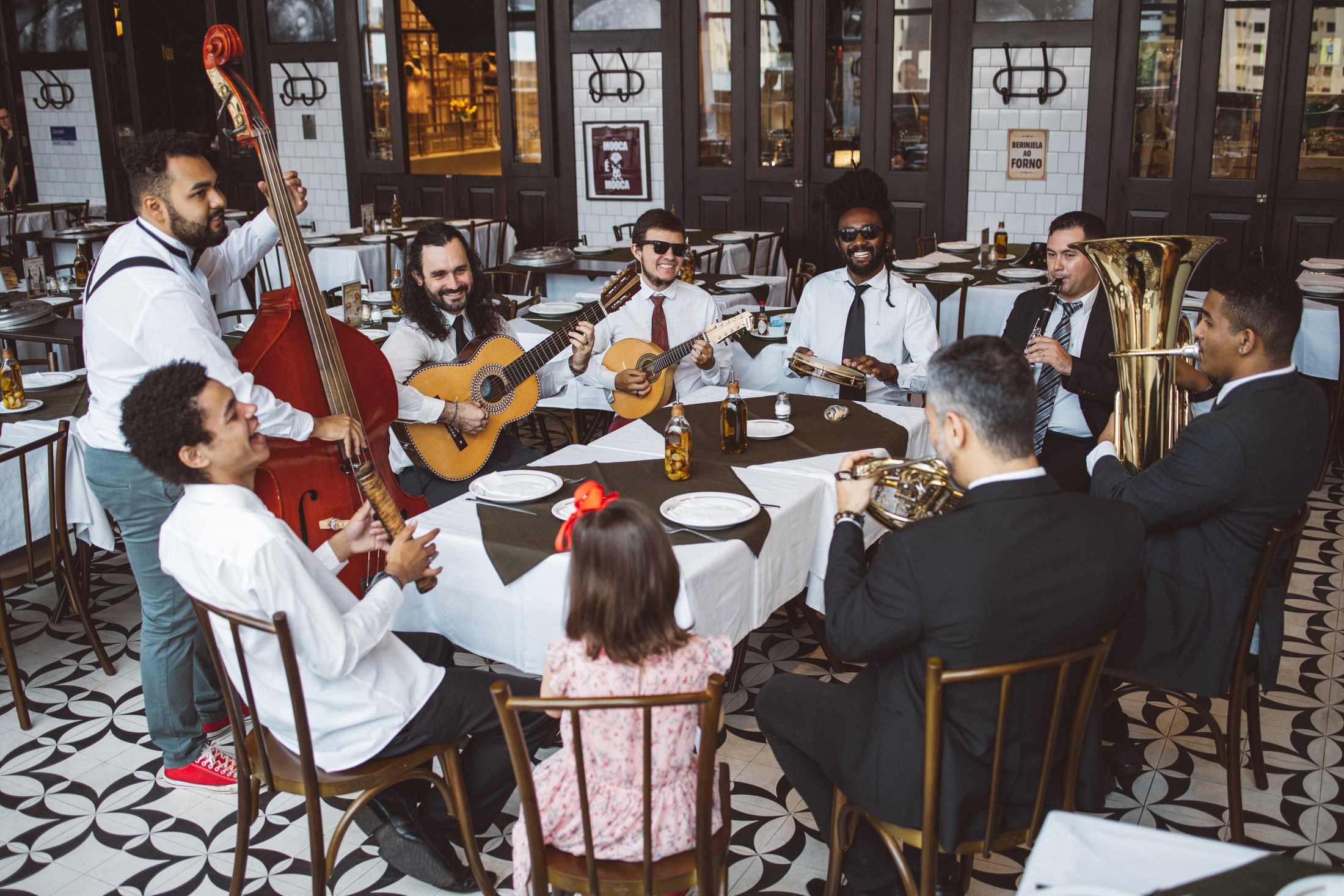 Regional do Clube do Choro tocando em volta de uma mesa de restaurante: contrabaixo, violão, cavaquinho, pandeiro, tuba e trompete